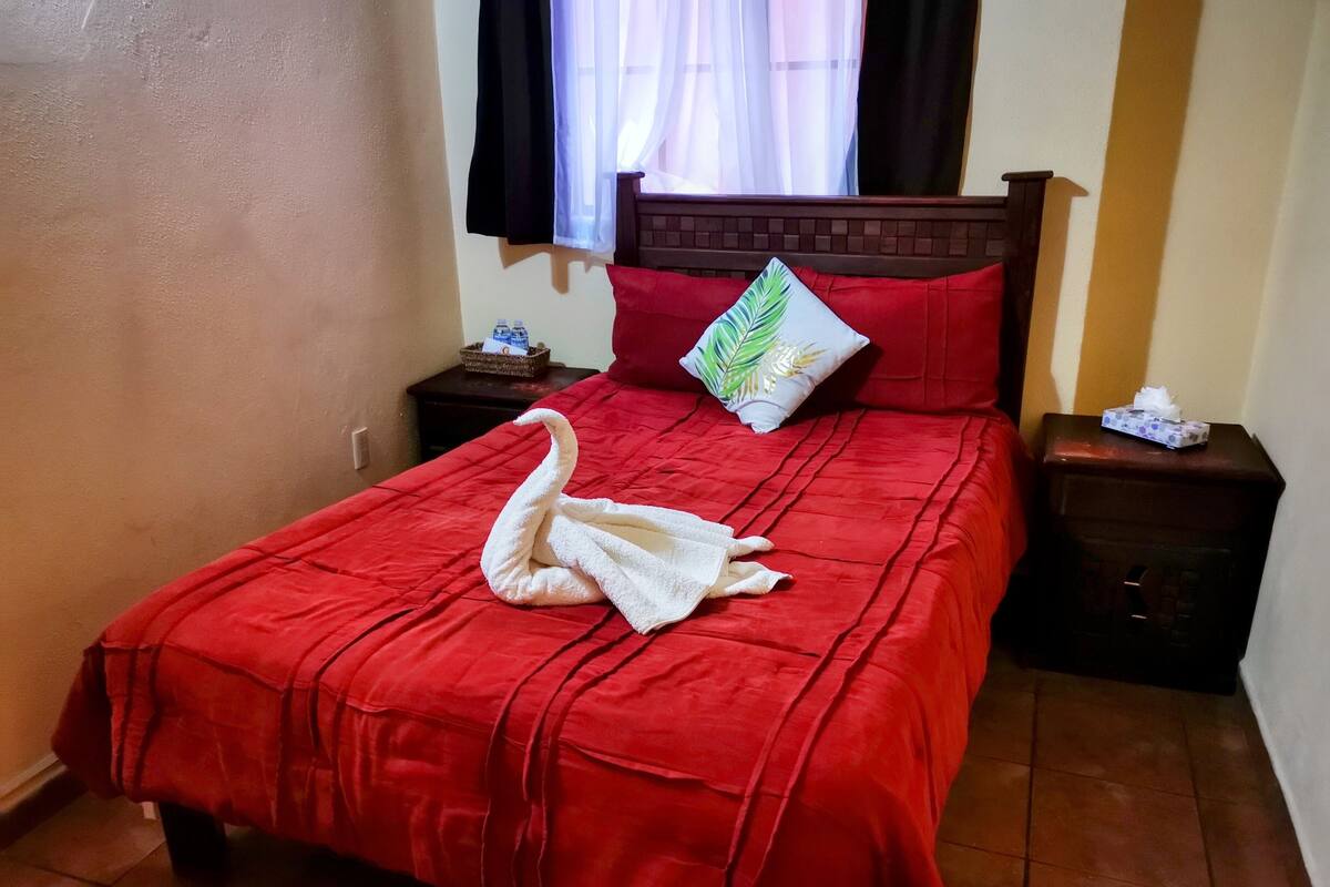 Una cama impecablemente hecha con una colcha roja y una toalla blanca dispuesta en forma de cisne. Hay un almohadón con diseño de hoja, una mesita de noche con una caja de pañuelos y una ventana con cortinas traslúcidas.