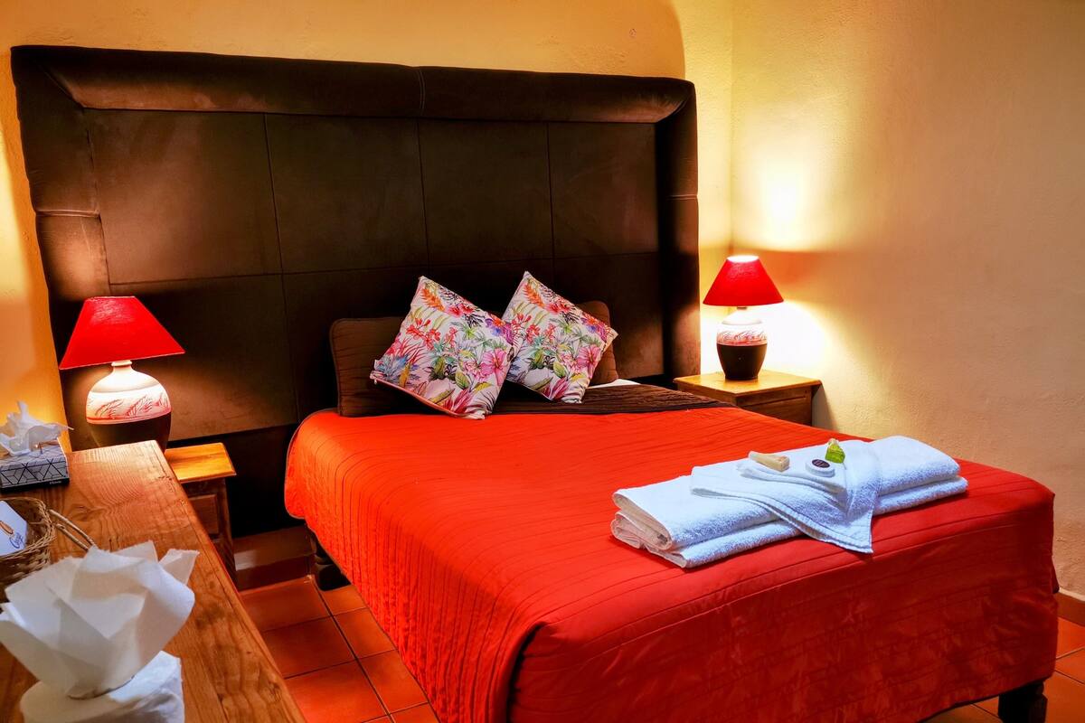 Una cama bien hecha con una colcha roja, dos almohadas con motivos florales y una toalla doblada en el pie. Hay dos mesitas de noche con lámparas, una de ellas con pantalla roja.