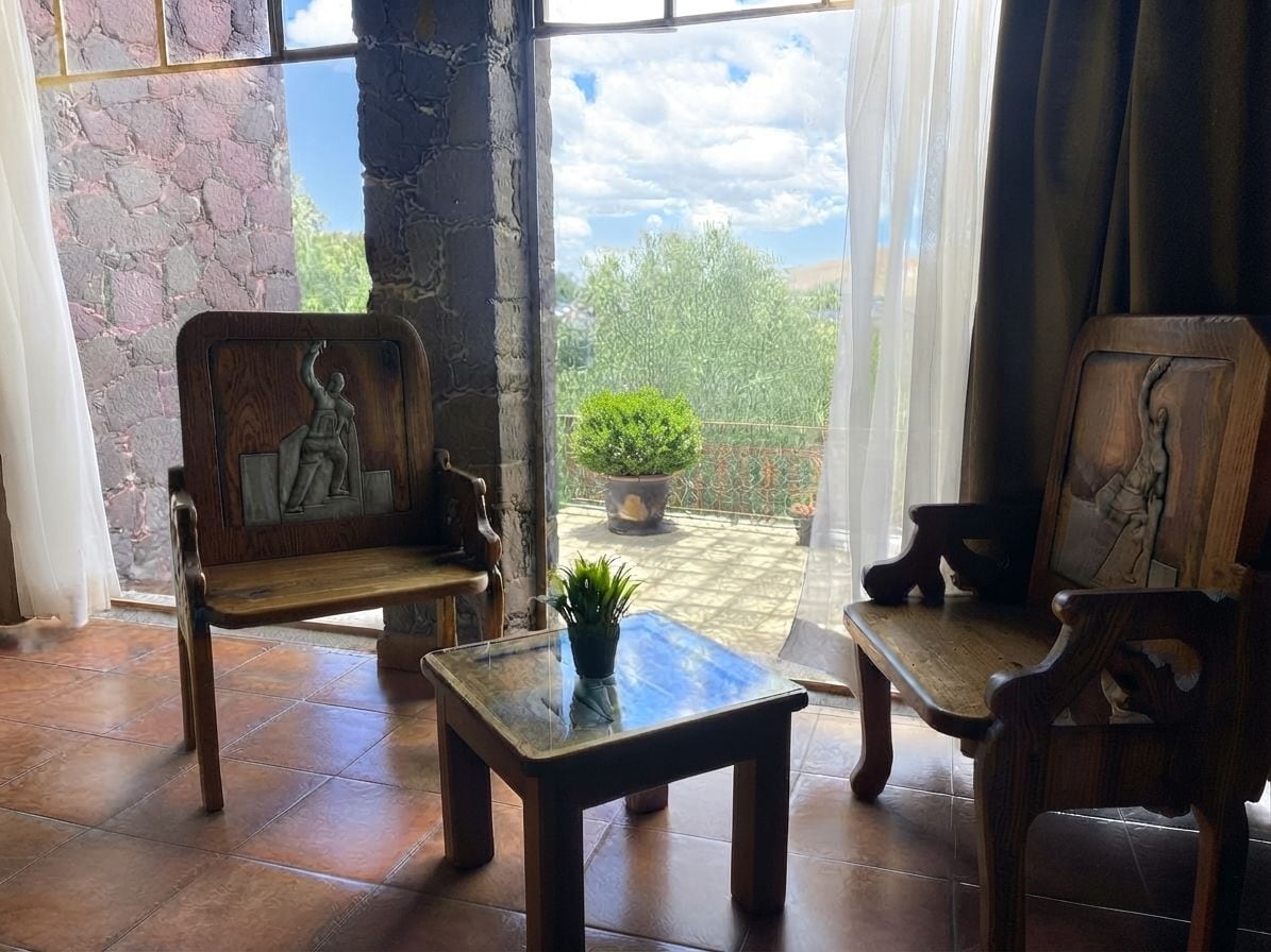 Una habitación con dos sillas de madera y una mesa pequeña, una planta en maceta, y vistas a un jardín a través de una ventana.