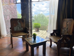 Una habitación con dos sillas de madera y una mesa pequeña, una planta en maceta, y vistas a un jardín a través de una ventana.