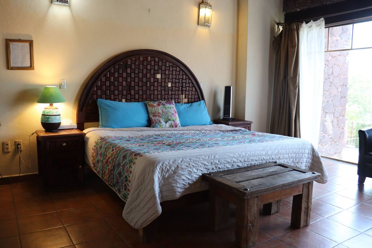 Un dormitorio con una cama grande, un banco de madera, una mesita de noche con una lámpara y un televisor montado en la pared.