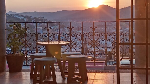 Un balcón con una mesa y sillas, una planta en maceta y vistas de un paisaje urbano con montañas al fondo.