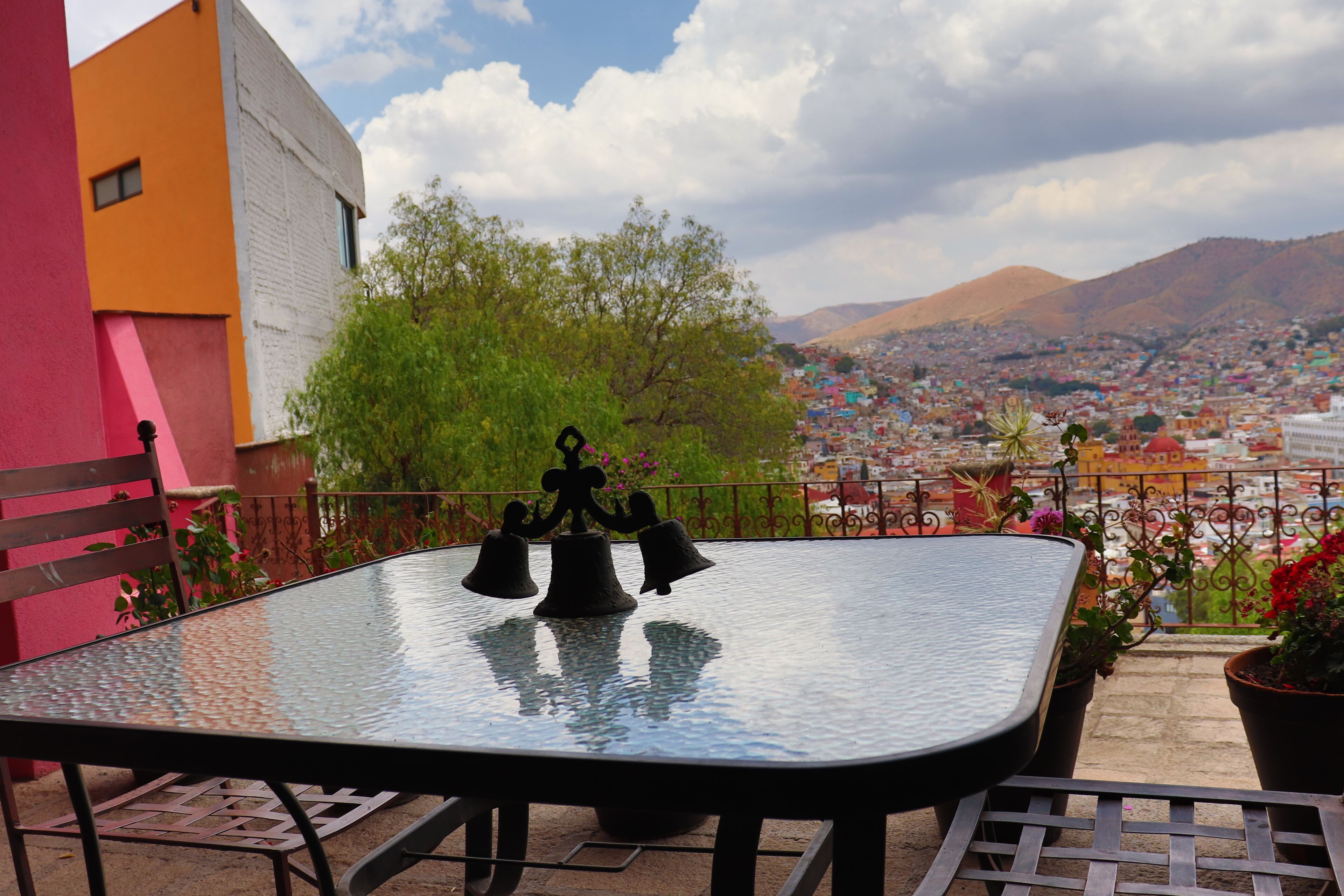 Una mesa con cubierta de vidrio y sillas de metal en un balcón con vistas a un paisaje urbano y montañas al fondo.