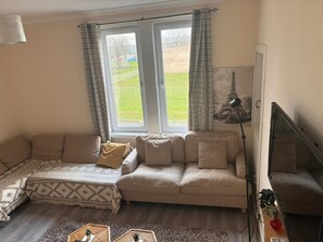 A living room with a sofa, a small table with a plant, a lamp, and a view of a park through the window. - Beach & City Access Flat 2 Floors, Full Kitchen near train 7mins (Methil)