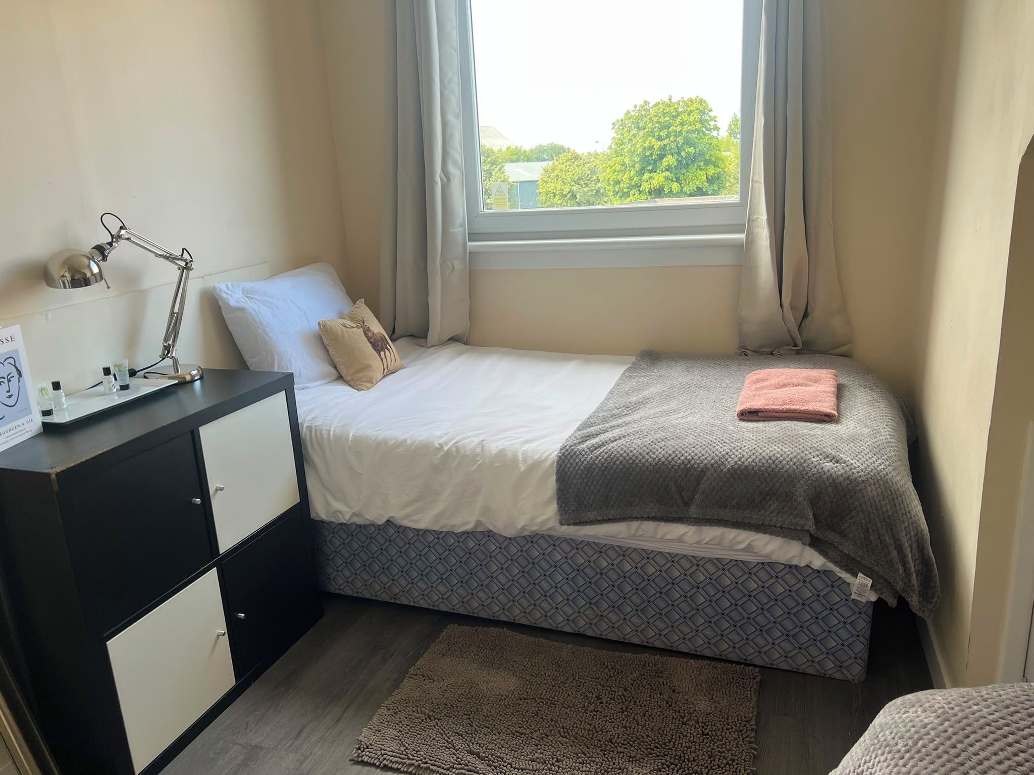Un dormitorio pequeño y ordenado con cama, mesita de noche, lámpara y vistas al exterior.