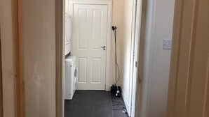 Een smalle gang met een witte deur aan het einde, een wasmachine en een stopcontact op de muur.