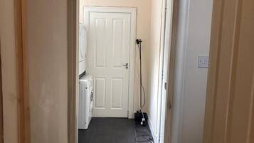 Een smalle gang met een witte deur aan het einde, een wasmachine en een stopcontact op de muur.