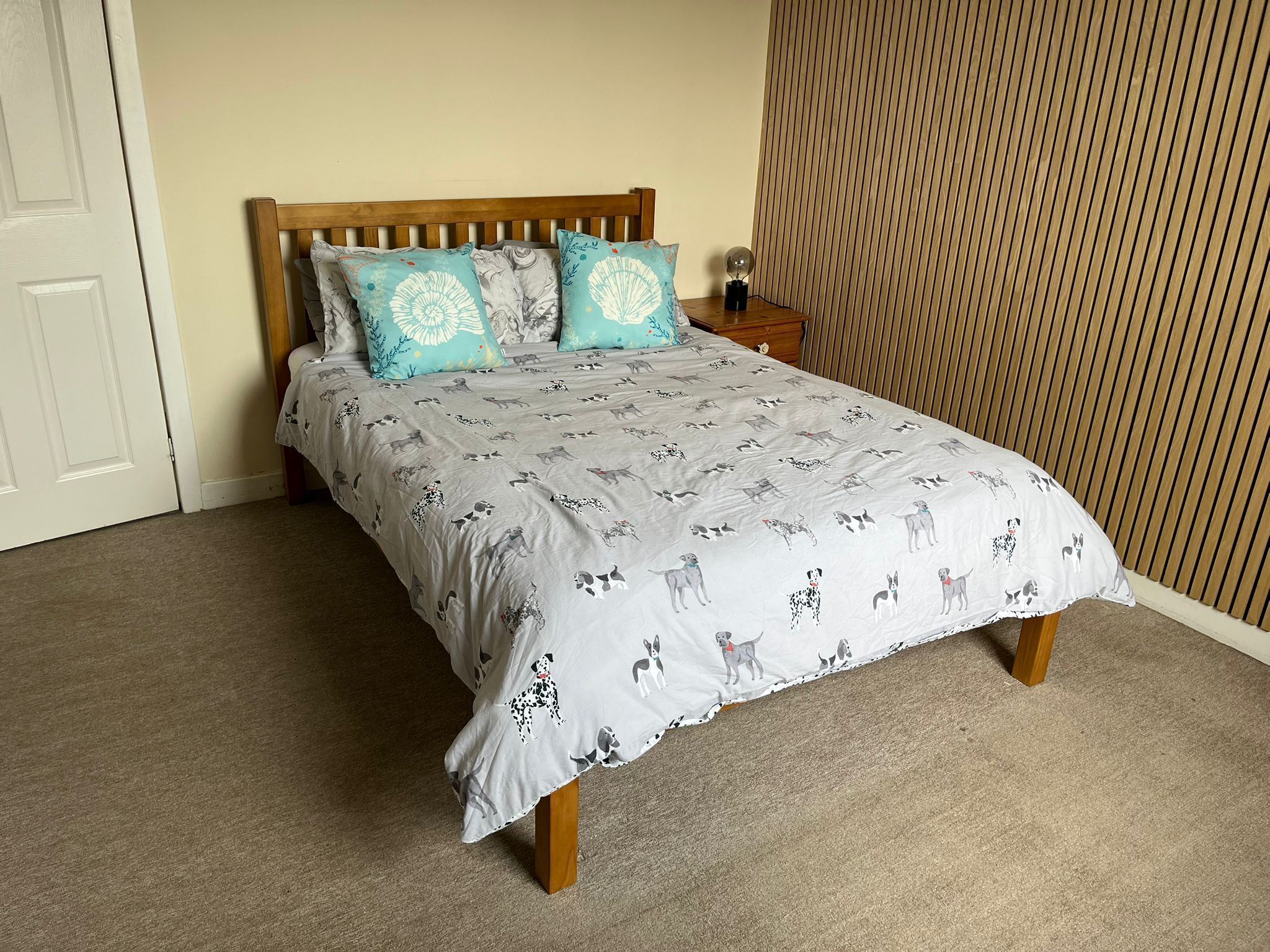 A bed with a dog print bedspread, two patterned pillows, a wooden headboard, and a nightstand with a lamp.