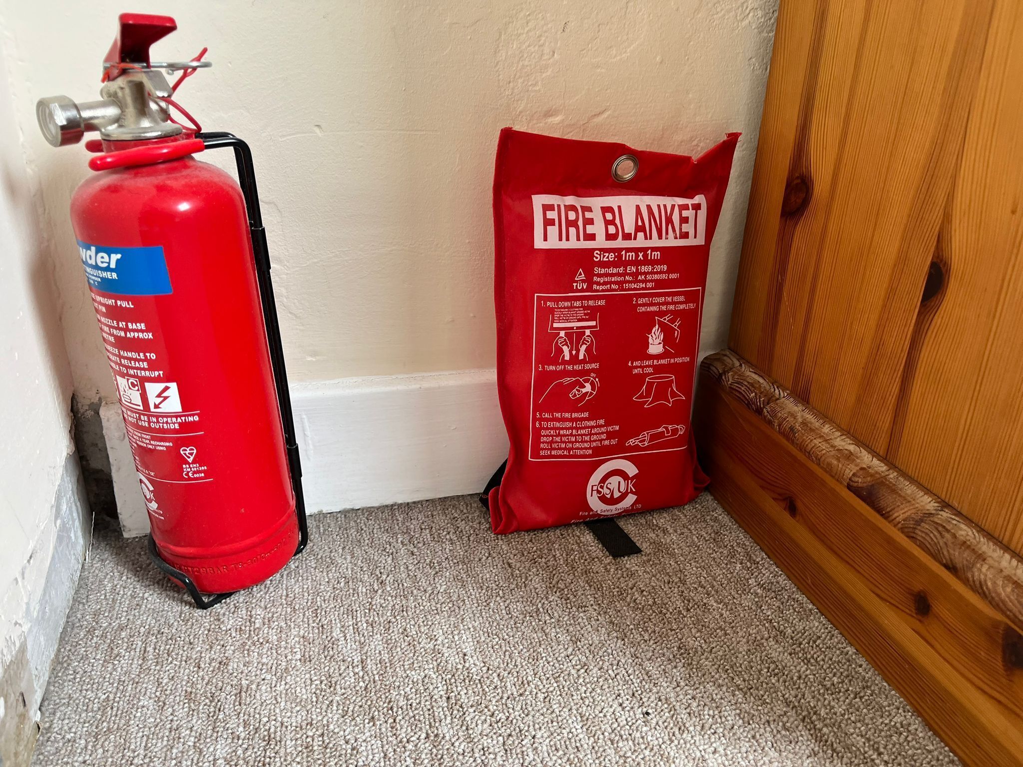 A fire extinguisher and a fire blanket on a carpeted floor next to a wooden door.