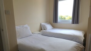 Une petite chambre propre, avec deux lits simples, une fenêtre avec des rideaux et une table de chevet blanche.