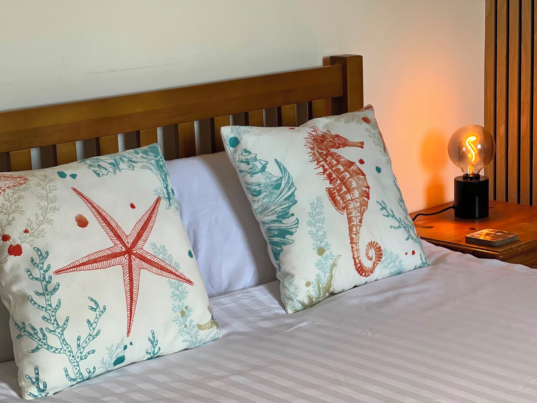Cama com duas almofadas decorativas com tema marinho, cabeceira de madeira e mesa de cabeceira com abajur.
