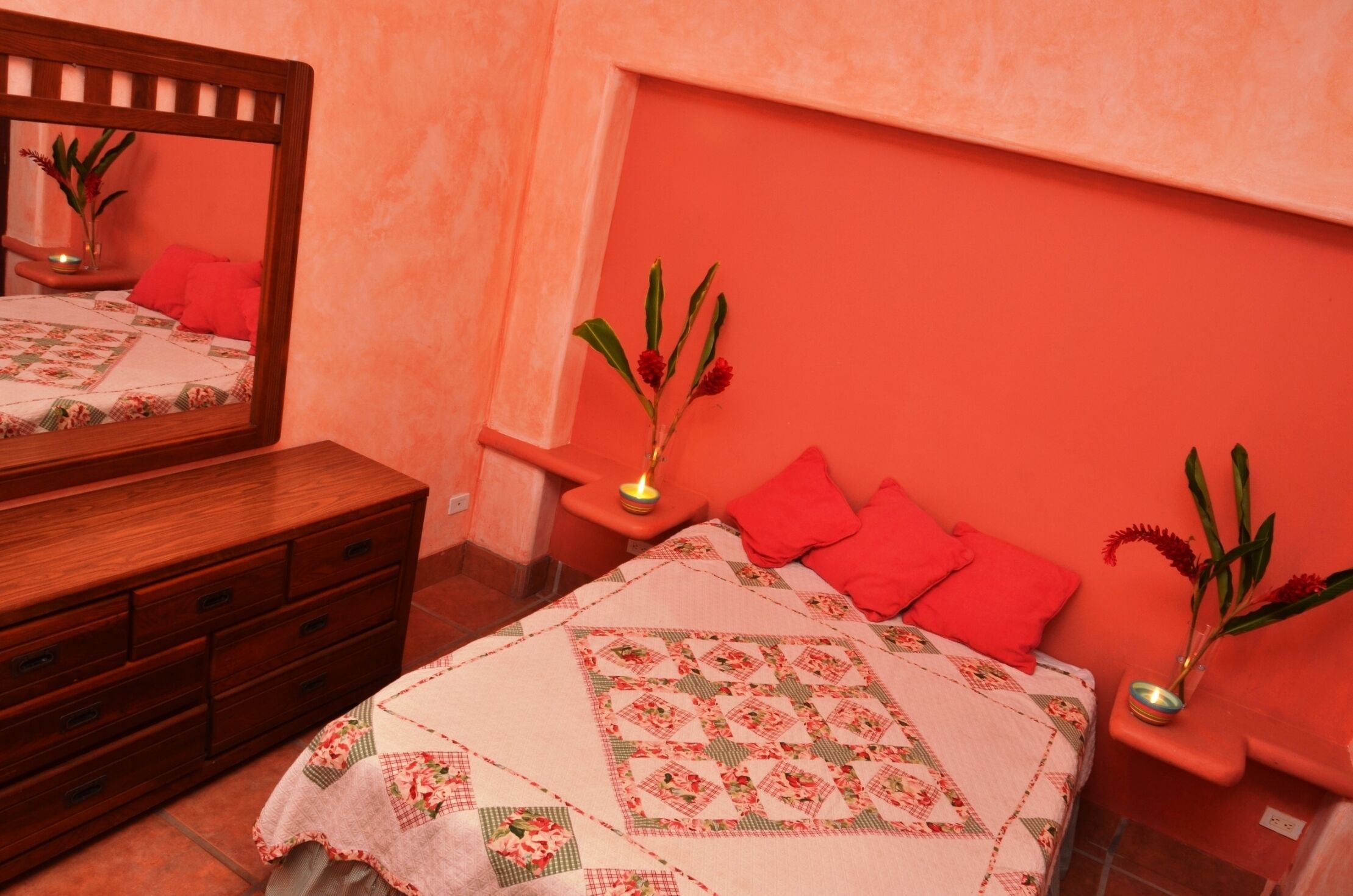 A bedroom with a wooden dresser, a bed with a floral quilt, two red pillows, and two bedside tables with candles and plants.