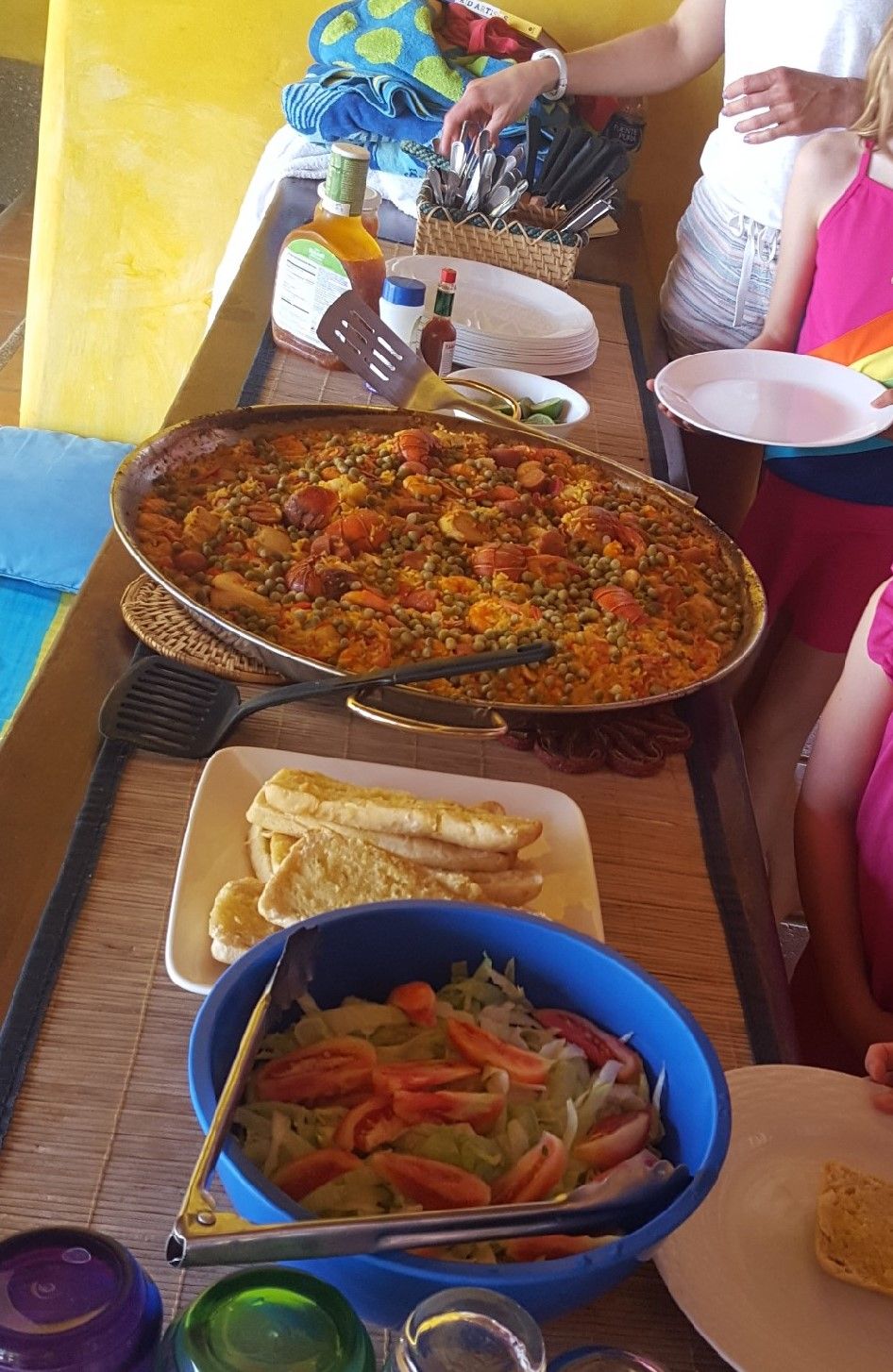 Una amplia mesa de comedor comunal con una paella, pan y un bol de ensalada.