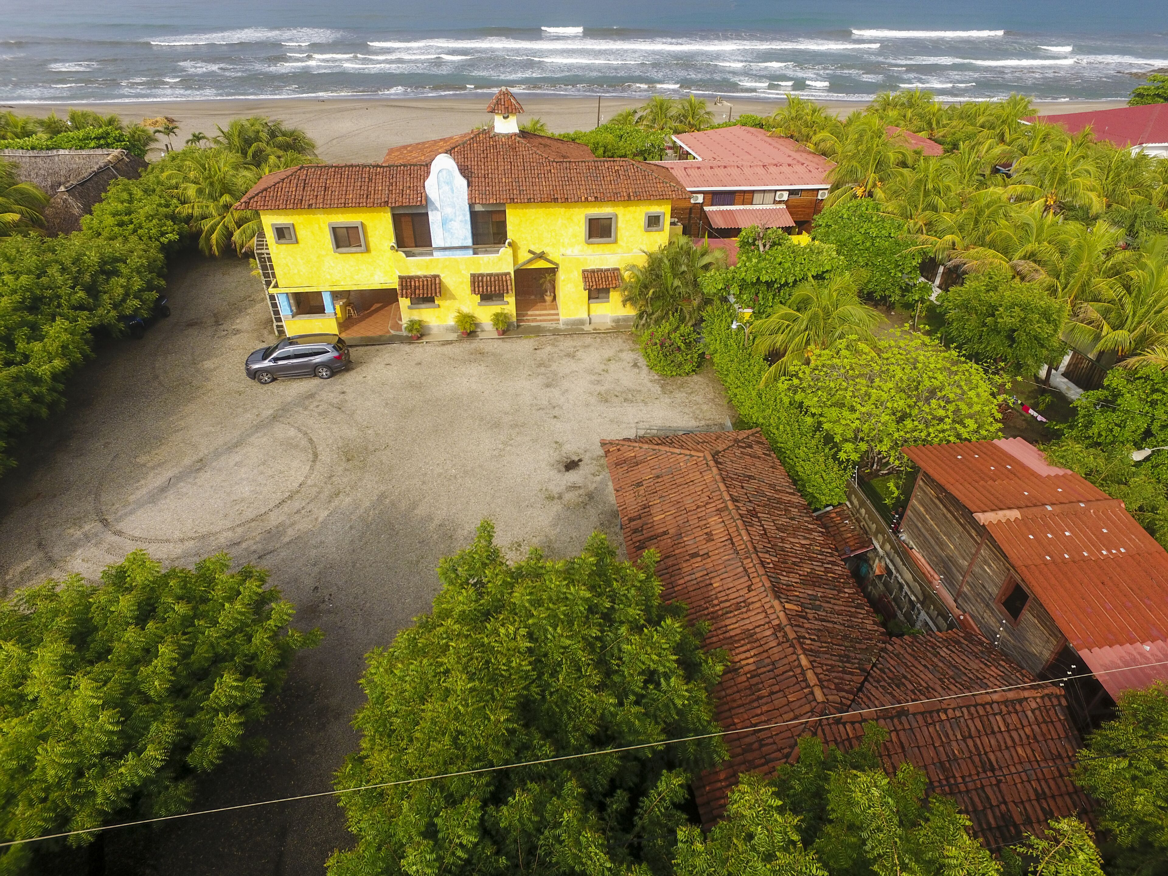 An aerial view of a yellow house with a red roof, surrounded by lush greenery and a beach in the background.