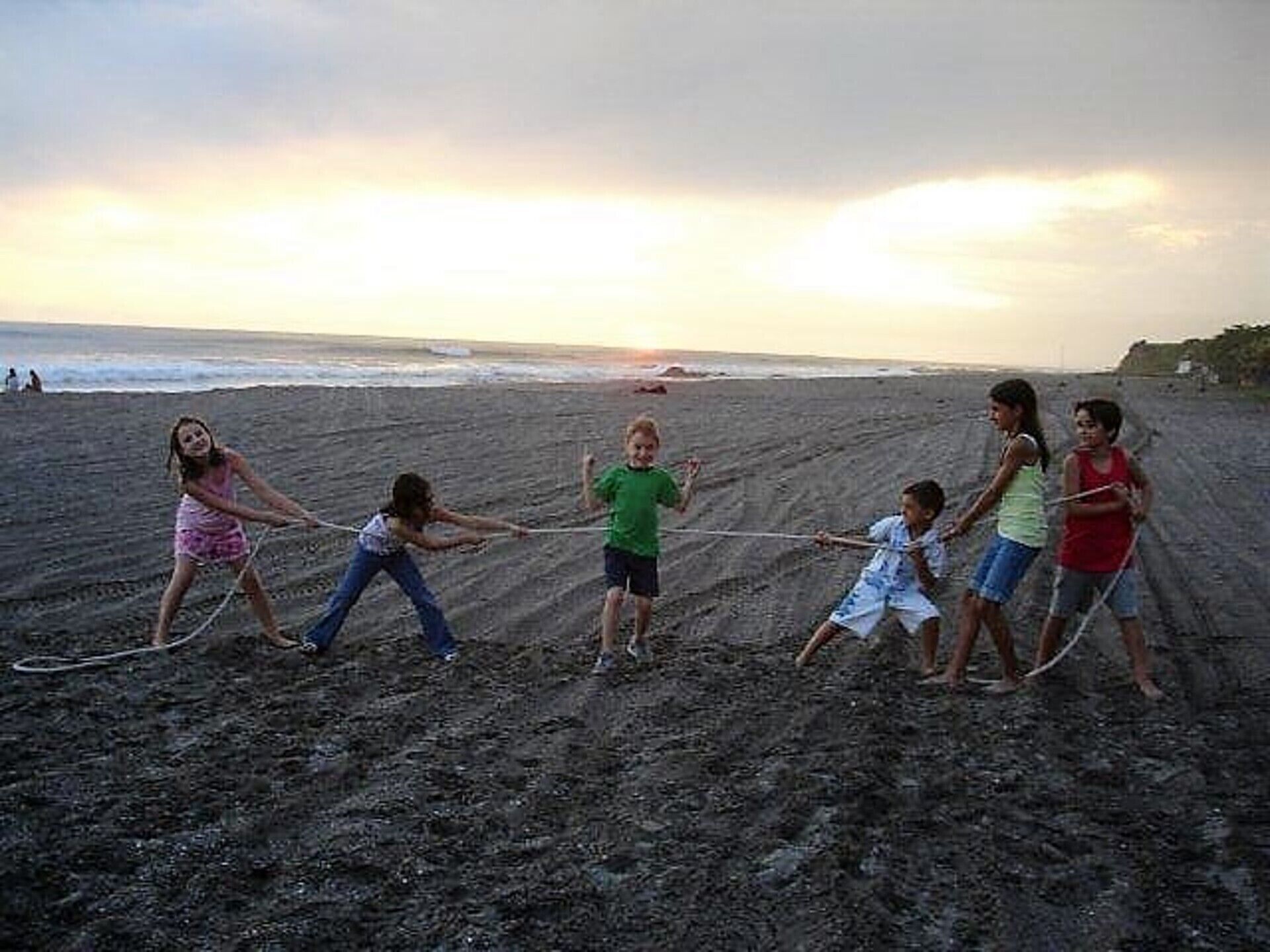 A group of children playing tug-of-war on a sandy beach with the ocean in the background.