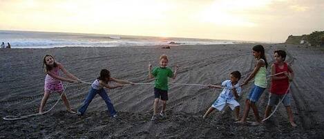 A group of children playing tug-of-war on a sandy beach with the ocean in the background.