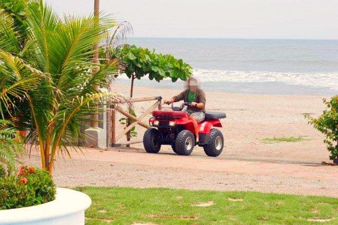 A person riding a red ATV on a beach path with palm trees and a view of the ocean.