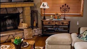 A cozy living room with a stone fireplace, a wooden coffee table, a sofa, and a window with blinds. - Lovely Cabin with lake views (Lake Arrowhead)