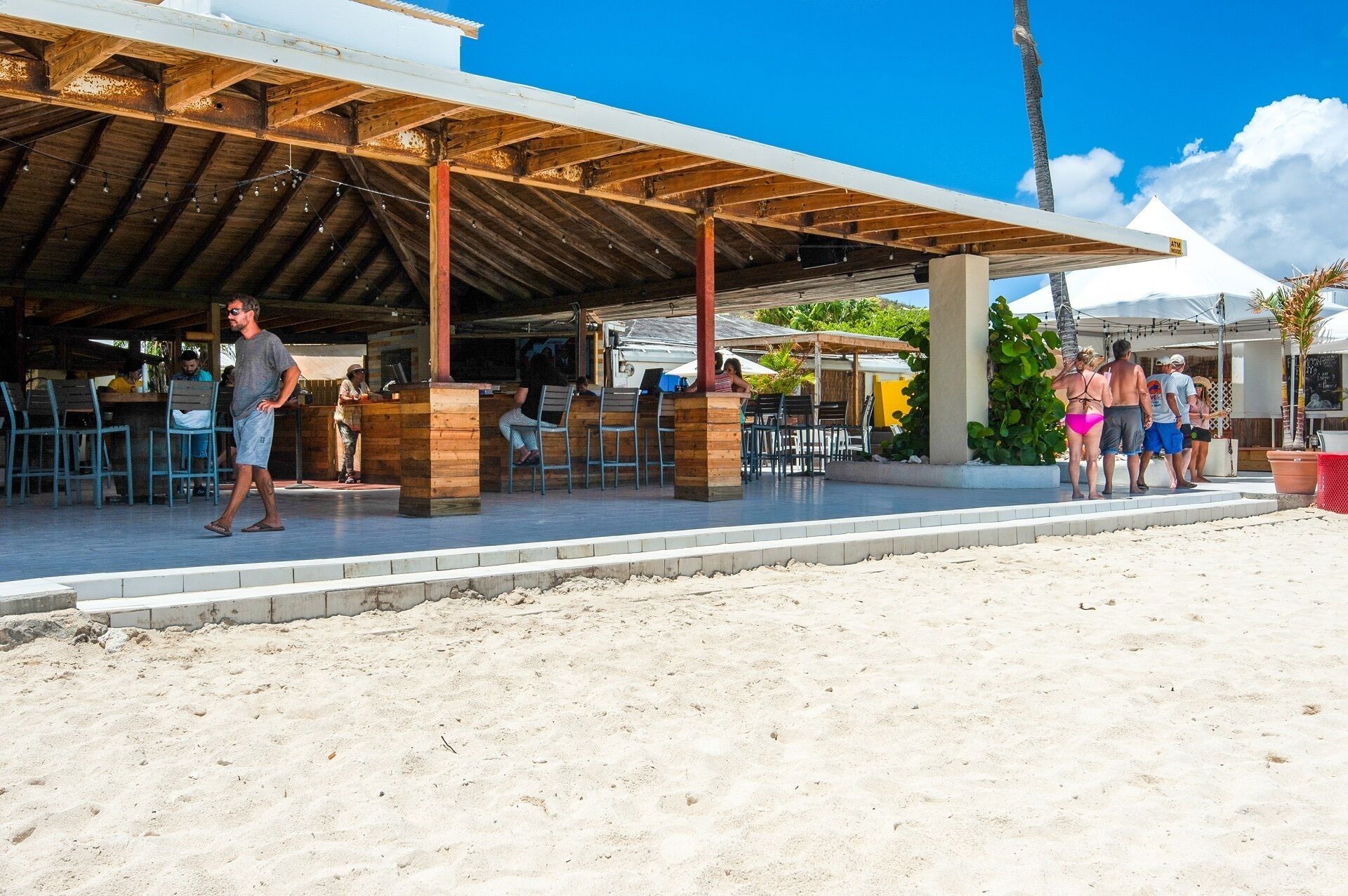 A beachside restaurant with outdoor seating, a covered area, and a sandy beach in the foreground.