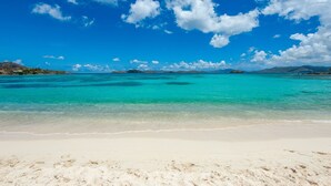 En strand med krystalklart turkis vand, hvid sand og fjerne bjerge under en blå himmel med skyer.