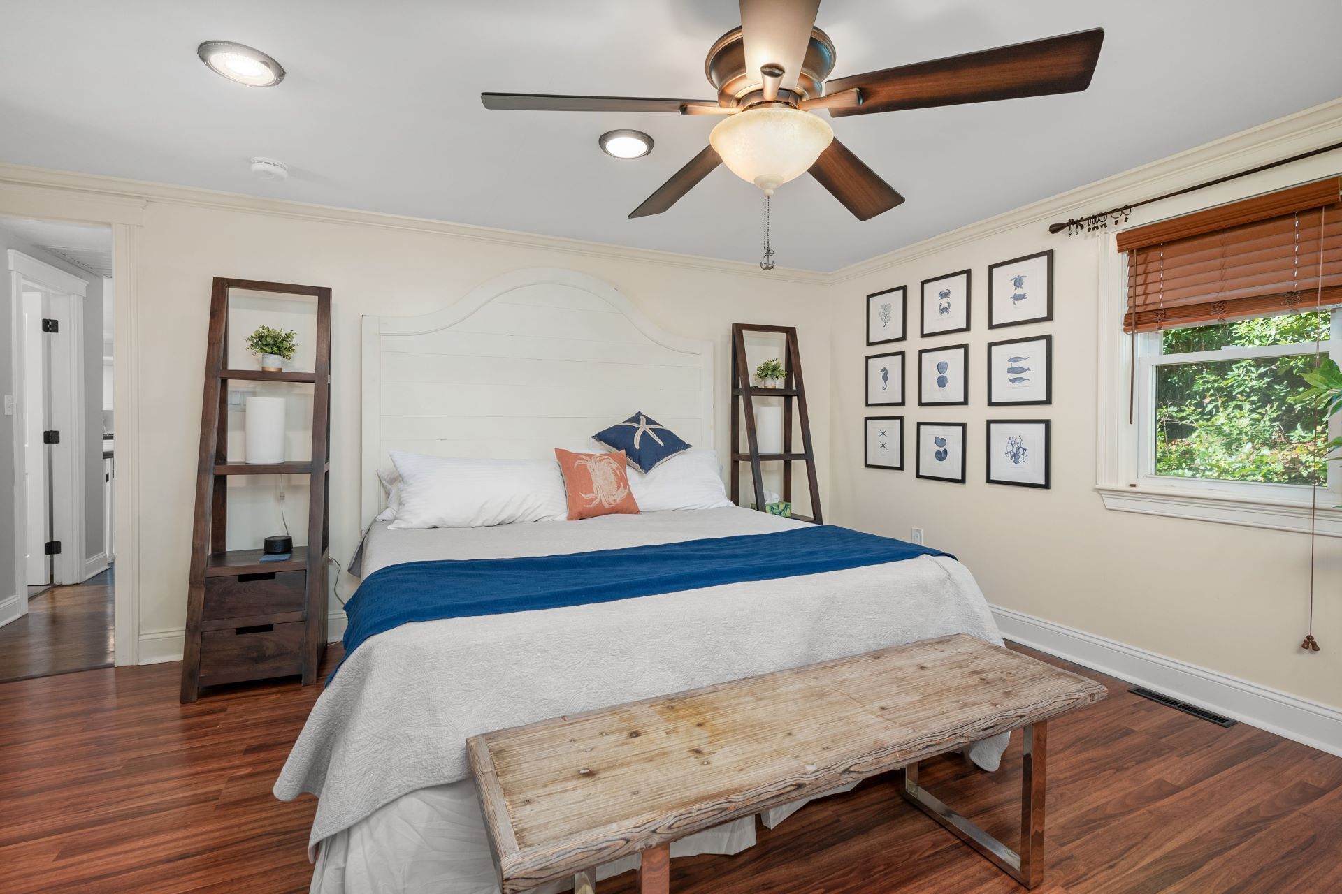 A bedroom with a bed, wooden bench, ceiling fan, framed pictures, and a window with blinds.