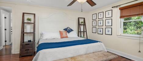 A bedroom with a bed, wooden bench, ceiling fan, framed pictures, and a window with blinds.