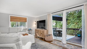 Ein modernes Wohnzimmer mit einer Couch, einem Fernseher und Blick ins Freie durch große Schiebeglasfenster.