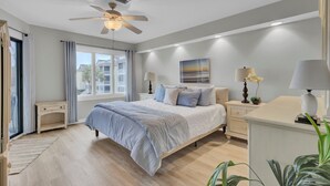 Une chambre à coucher avec un lit, un ventilateur de plafond, une fenêtre avec des rideaux, une table de chevet, une lampe et un tableau au mur.