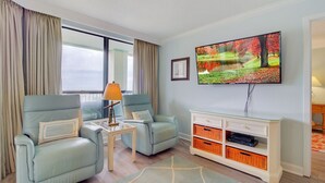 Un salon avec deux fauteuils bleu clair, une petite table avec une lampe, un téléviseur fixé au mur et un meuble de rangement blanc avec des paniers.