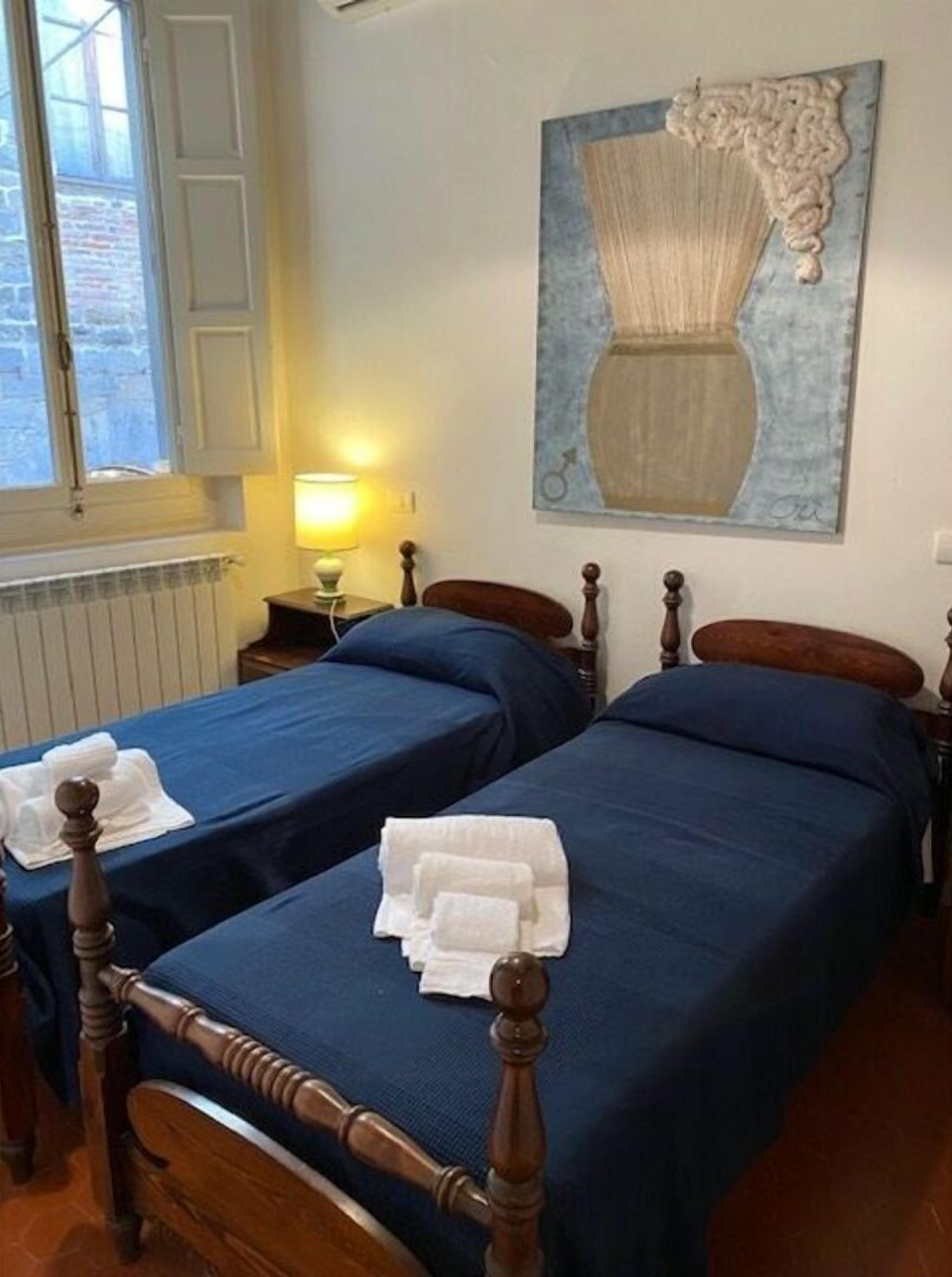 A hotel room with two wooden beds, each with a blue bedspread and white towels. There is a window with a view of a brick building, a lamp on the nightstand, and a painting on the wall.