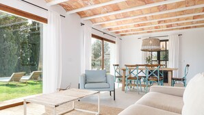 Amplia sala de estar con techo de madera, una mesa de comedor con sillas, un sofá y vista a un jardín a través de grandes ventanales.
