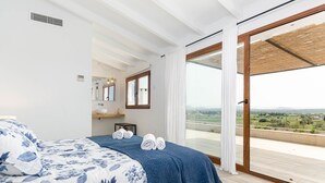 Un dormitorio con una cama grande, vistas al campo a través de un ventanal corredero y un baño pequeño con espejo y estante.