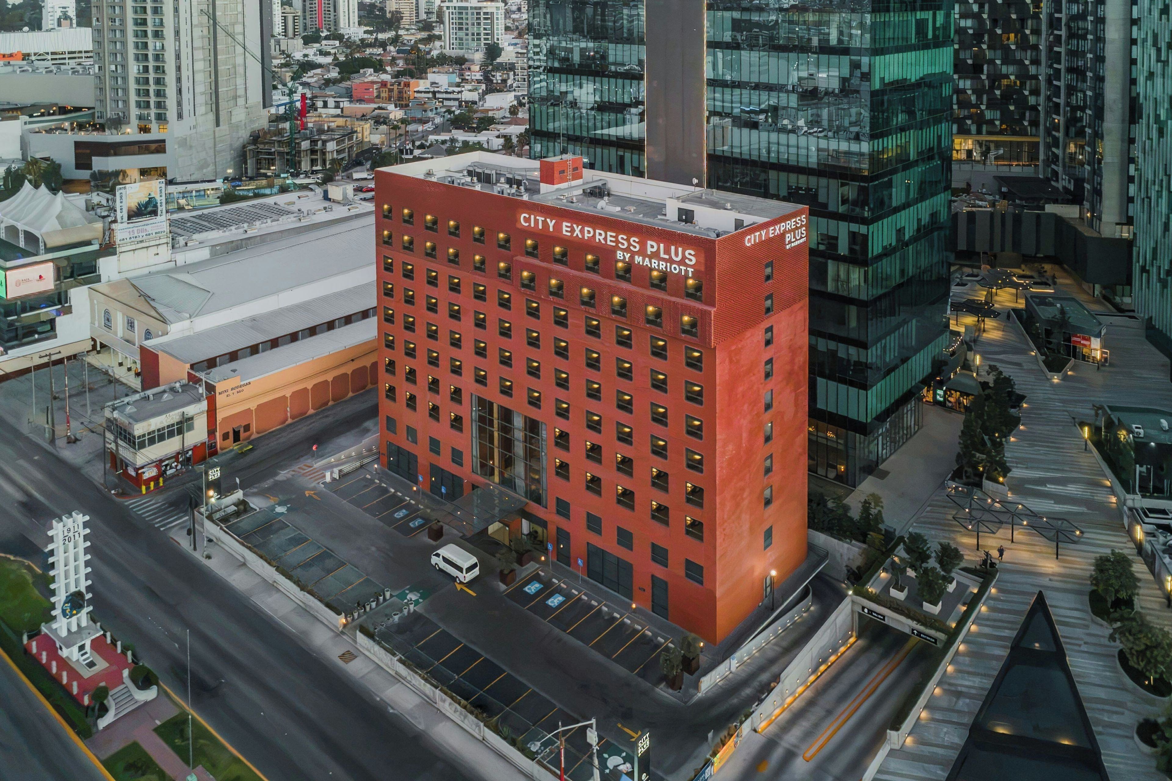 Un hotel de varios pisos con fachada roja, estacionamiento y paisaje urbano al fondo.