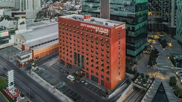 Un hotel de varios pisos con fachada roja, estacionamiento y paisaje urbano al fondo.