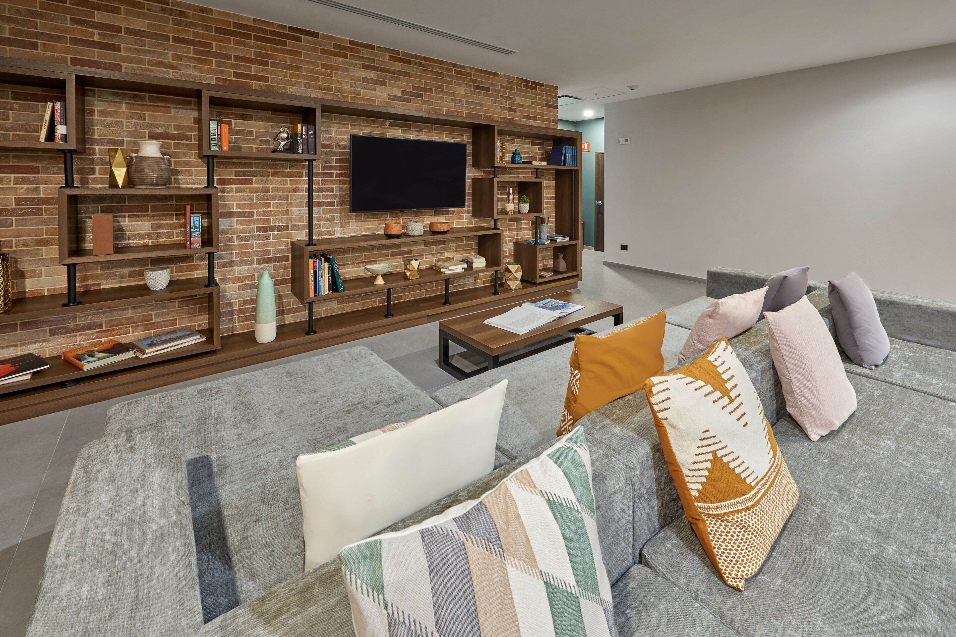 Uma sala de estar moderna com parede de tijolos, prateleiras embutidas, TV de tela plana e sofá cinza com almofadas estampadas.