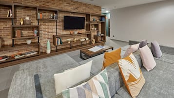 Uma sala de estar moderna com parede de tijolos, prateleiras embutidas, TV de tela plana e sofá cinza com almofadas estampadas.