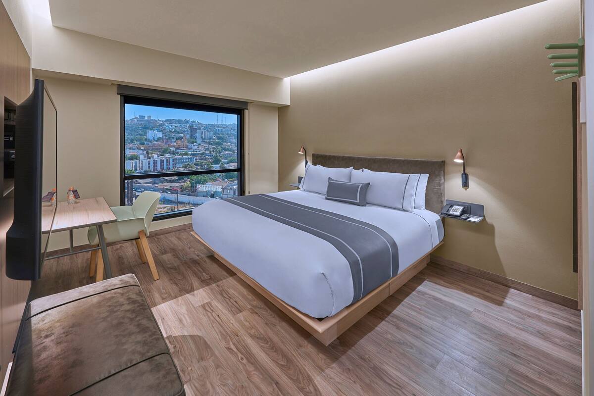 Habitación de hotel moderna con una cama grande, un escritorio, una silla y vistas a un paisaje urbano a través de un ventanal amplio.