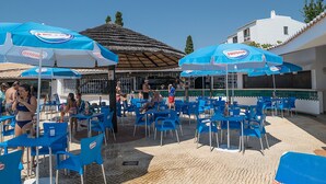 An outdoor seating area with blue chairs and tables, some with umbrellas, and a building with a tiled roof in the background. - Jasmine Apartment, AC, Pool, Balcony (Albufeira)