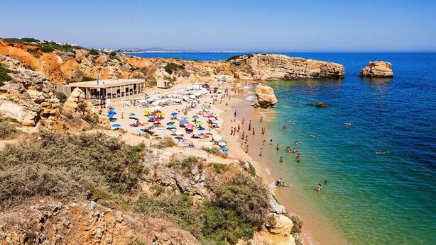 A beach with rocky terrain, clear blue water, and a building with a roofed area. - Jasmine Apartment, AC, Pool, Balcony (Albufeira)