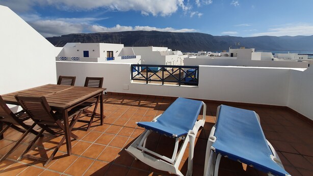 Una terraza con mesa y sillas, vista al mar y a las montañas, y edificios blancos con detalles azules.