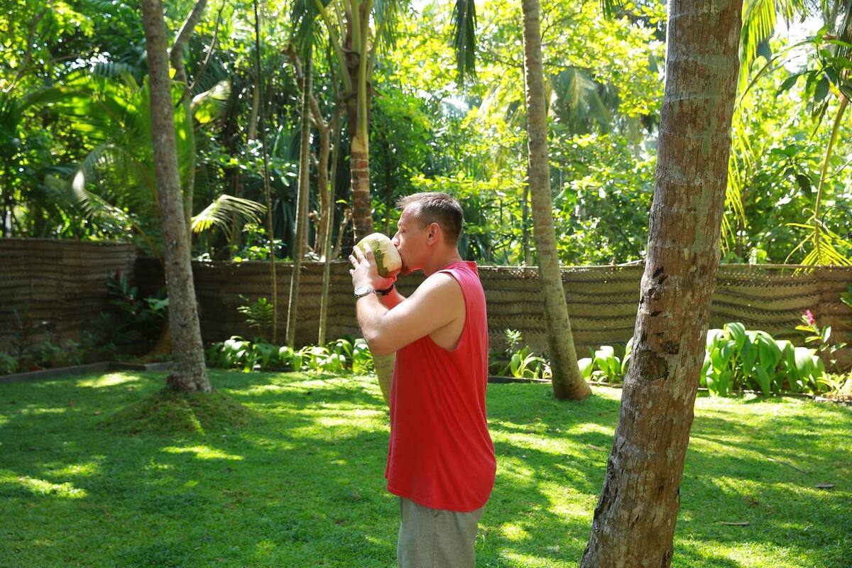 A man in a red shirt drinking from a coconut in a lush, green outdoor setting with trees and a wooden fence.