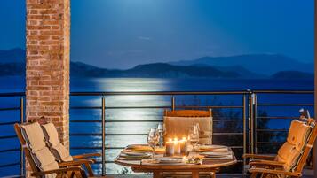 A dining table set for two with candles, glasses, and plates, overlooking a body of water with mountains in the background.