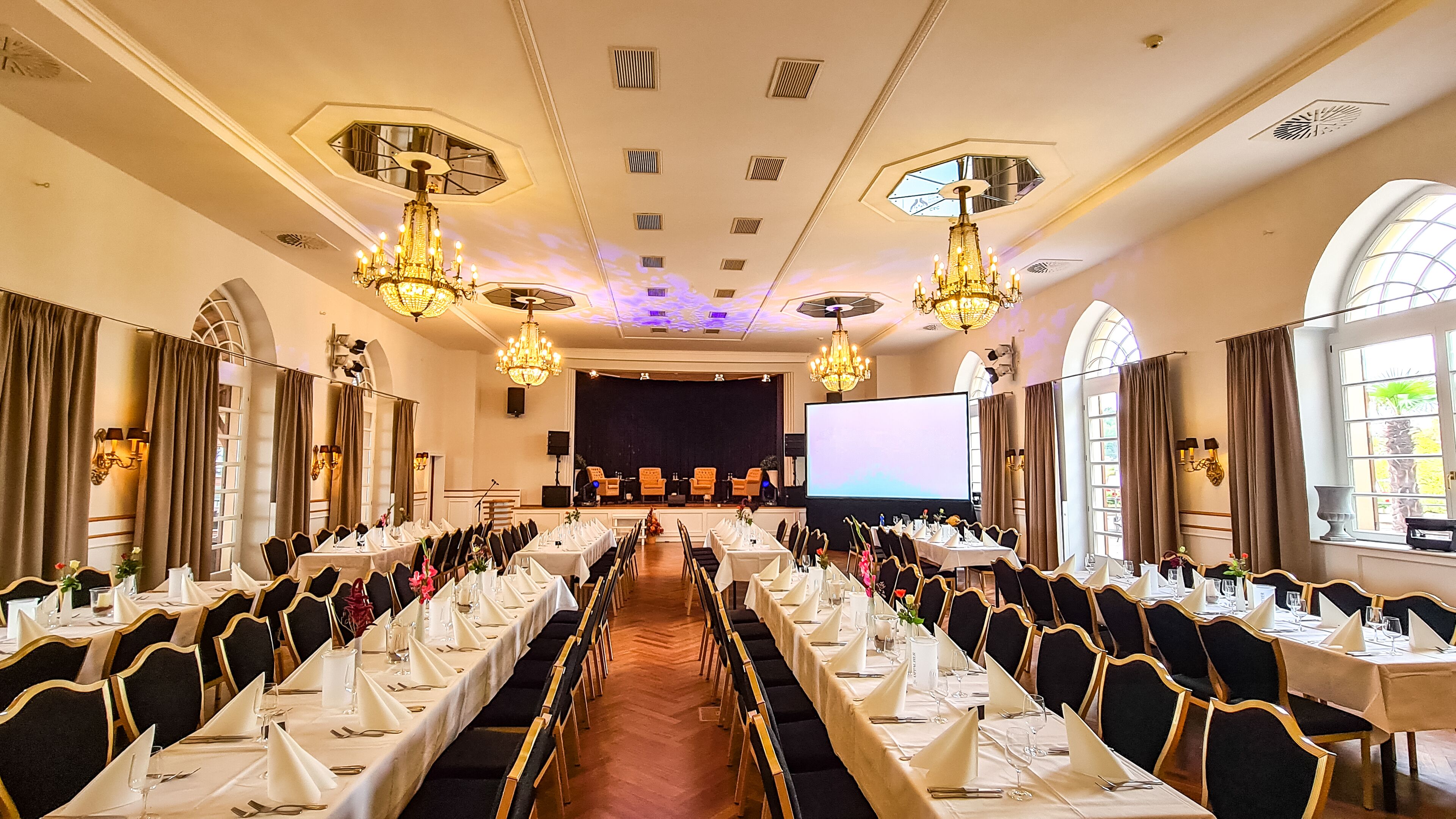 A conference room with long tables, black chairs, chandeliers, and a stage with a screen.