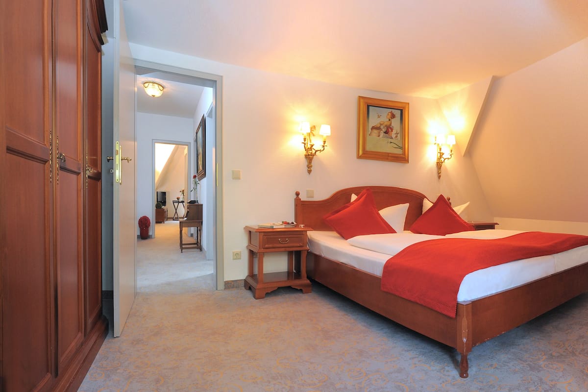 A bedroom with a large bed, two bedside lamps, a nightstand, a wooden wardrobe, and a painting on the wall.