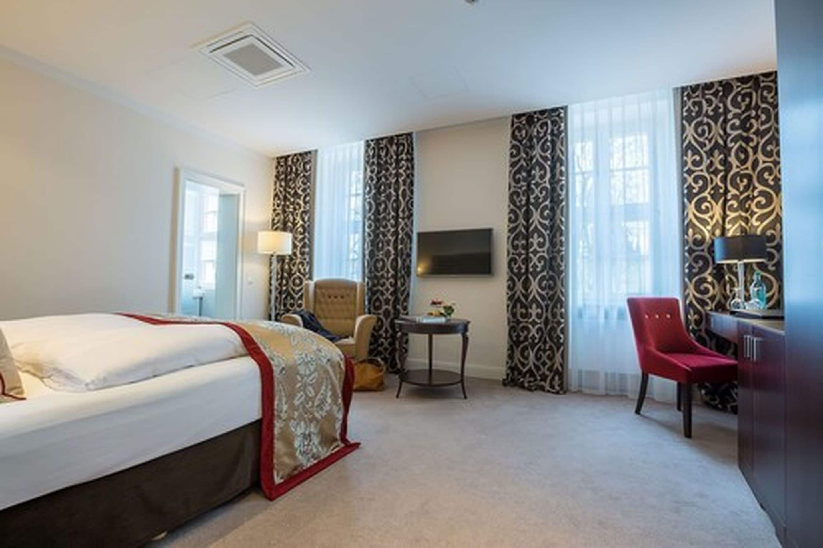 A hotel room with a bed, a desk, a chair, a TV, and curtains with a pattern.