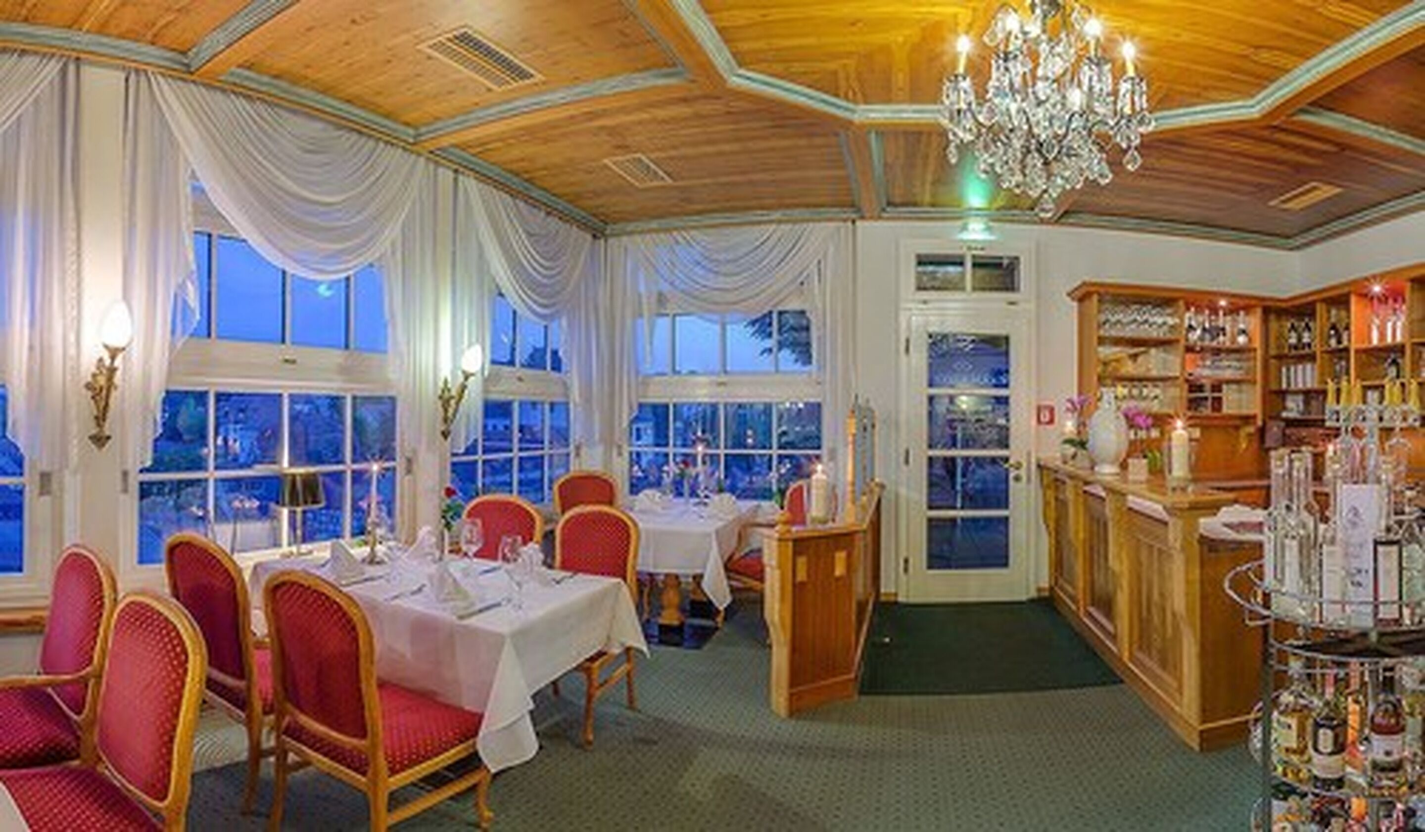 Un comedor con mesas dispuestas para cuatro, una zona de bar con estantes llenos de botellas y ventanas amplias con cortinas.