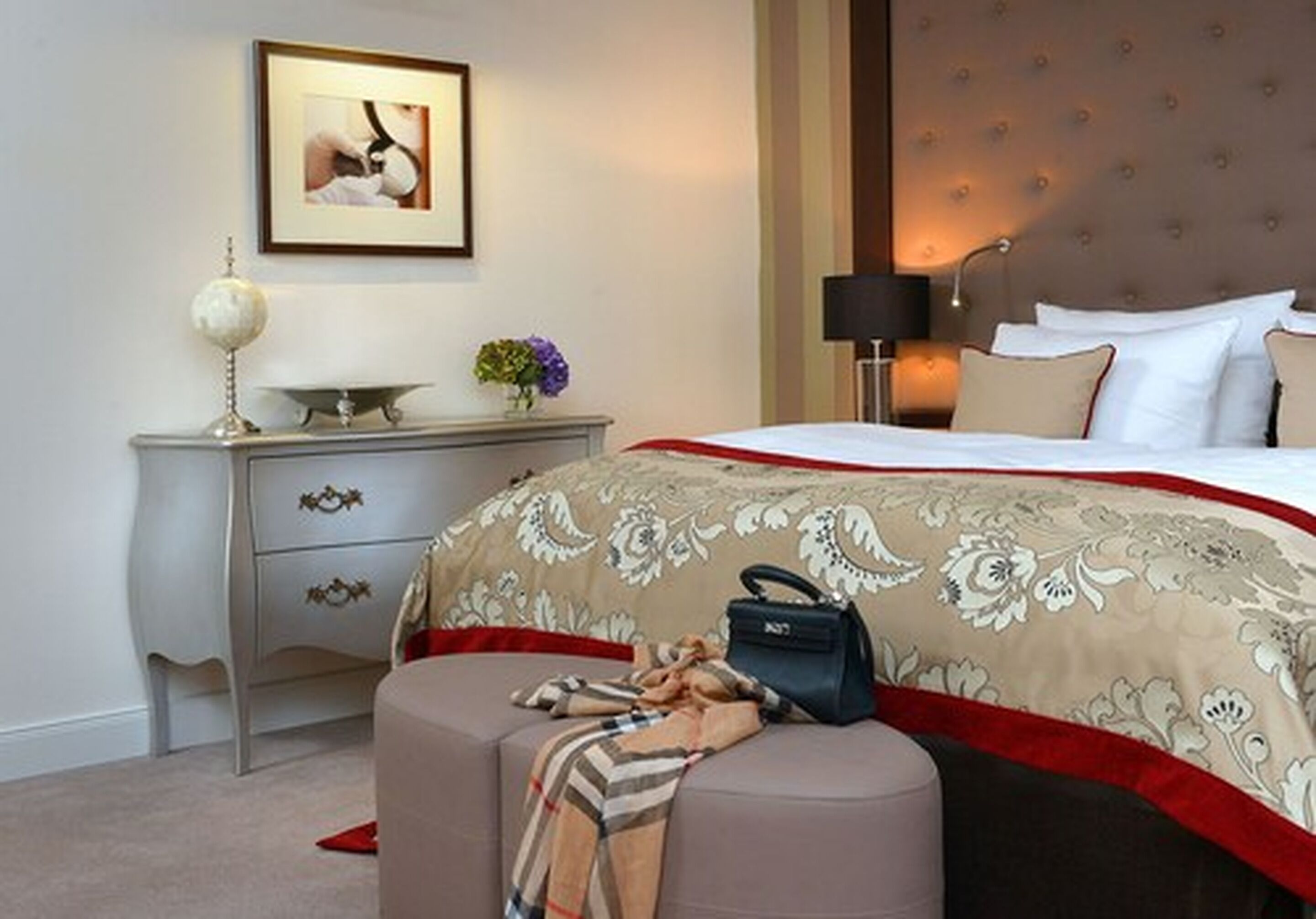 A hotel room with a bed, a nightstand, a lamp, a framed picture, and a small table with a globe and flowers.