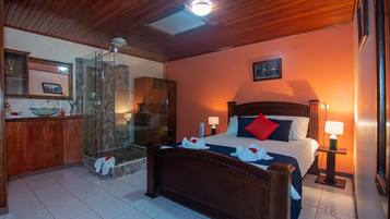 Une chambre d’hôtel avec un plafond en bois, un lit avec des oreillers rouges et blancs, une table de chevet avec une lampe, une salle de bain avec une douche en verre et un meuble en bois avec un lavabo.