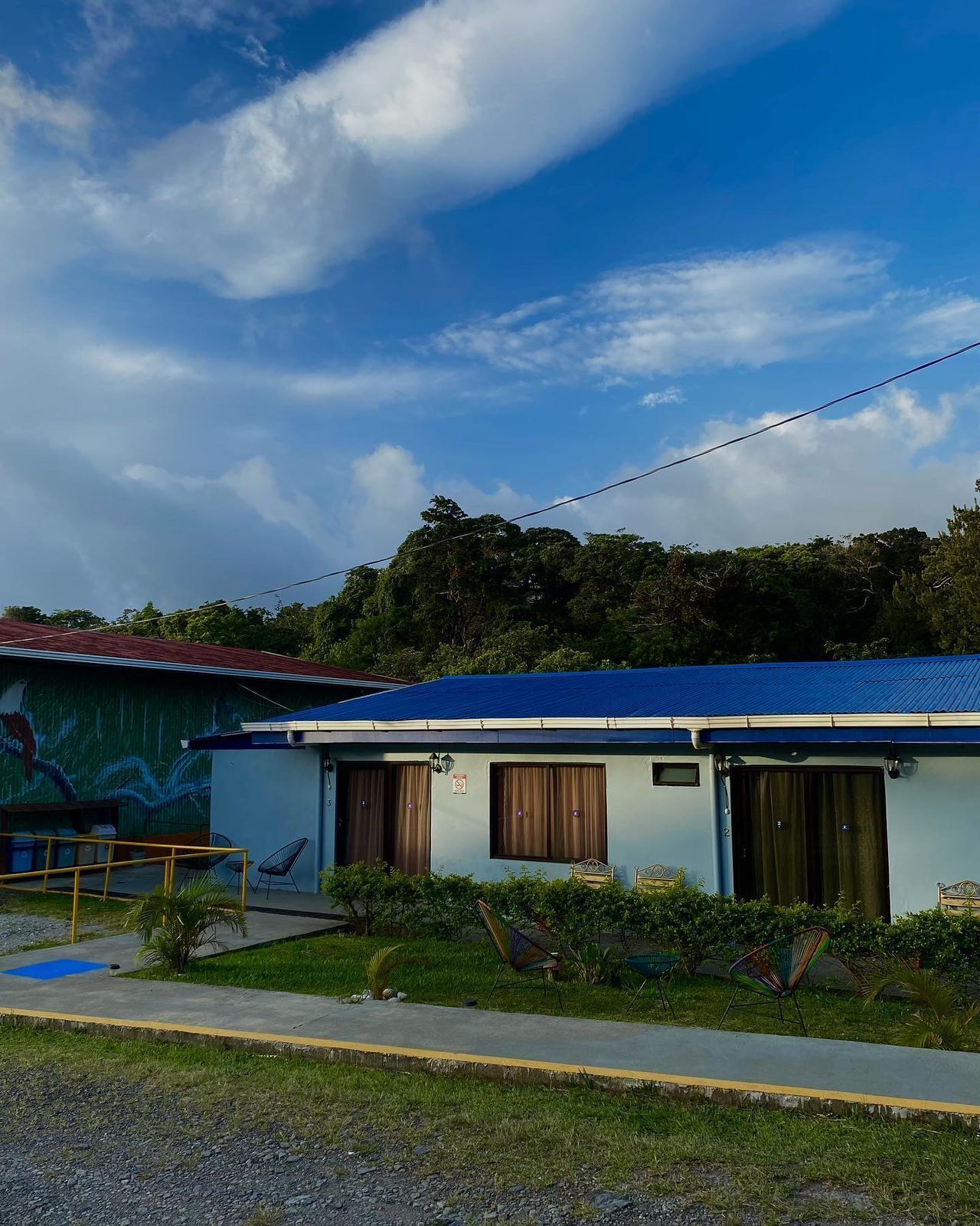 Un bâtiment à deux étages, avec un toit bleu, une fresque sur le côté et un petit jardin à l’avant.