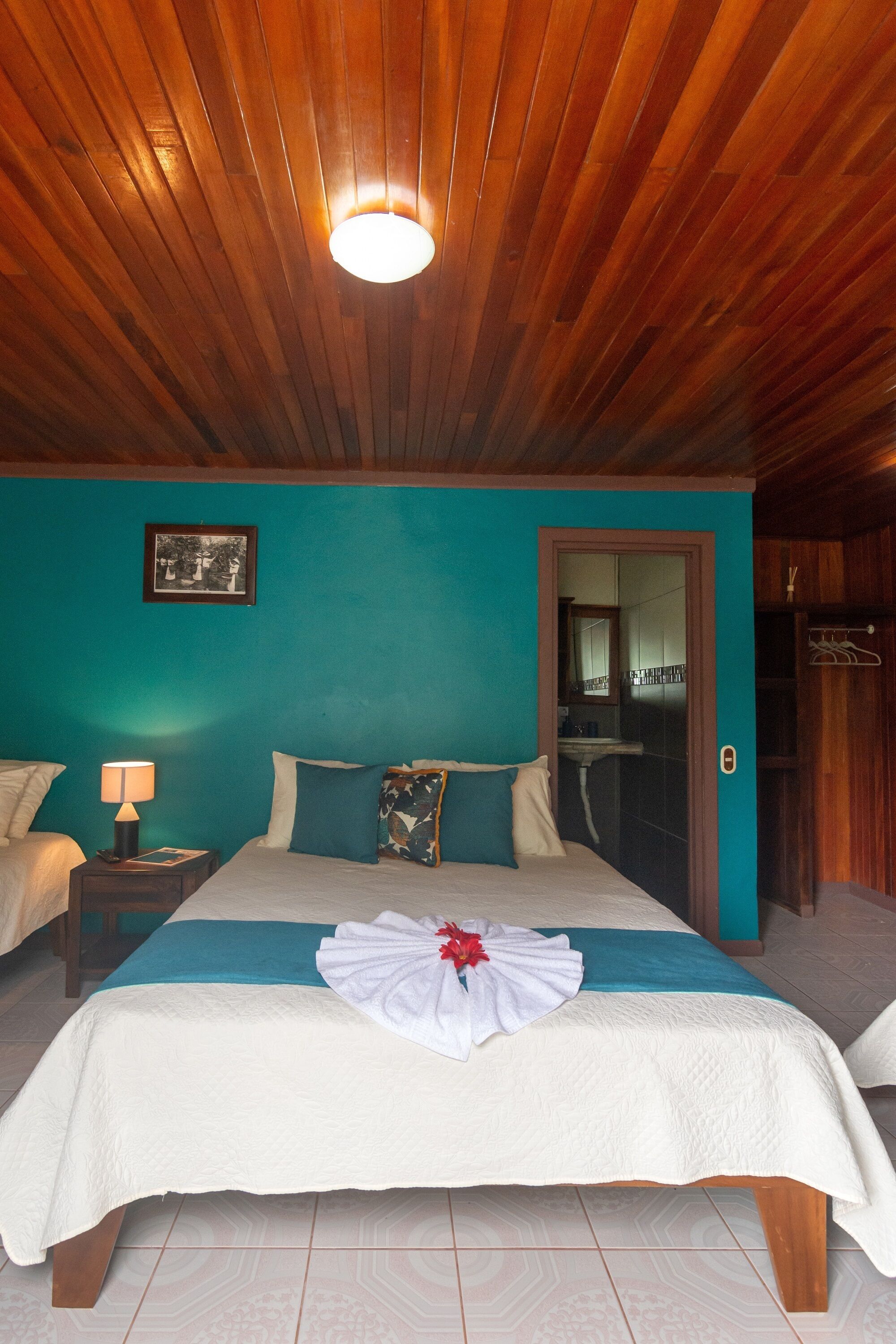 A hotel room with a wooden ceiling, teal walls, a bed with a white and teal bedspread, a bedside table with a lamp, and a bathroom visible through an open door.